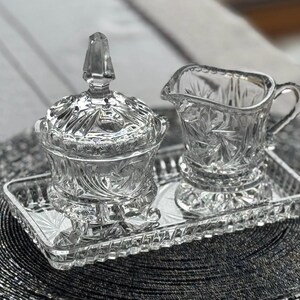 May include: A clear glass creamer and a lidded sugar bowl with a faceted finial, both featuring intricate starburst and geometric patterns, arranged together on a matching rectangular glass tray with a textured, scalloped edge design.