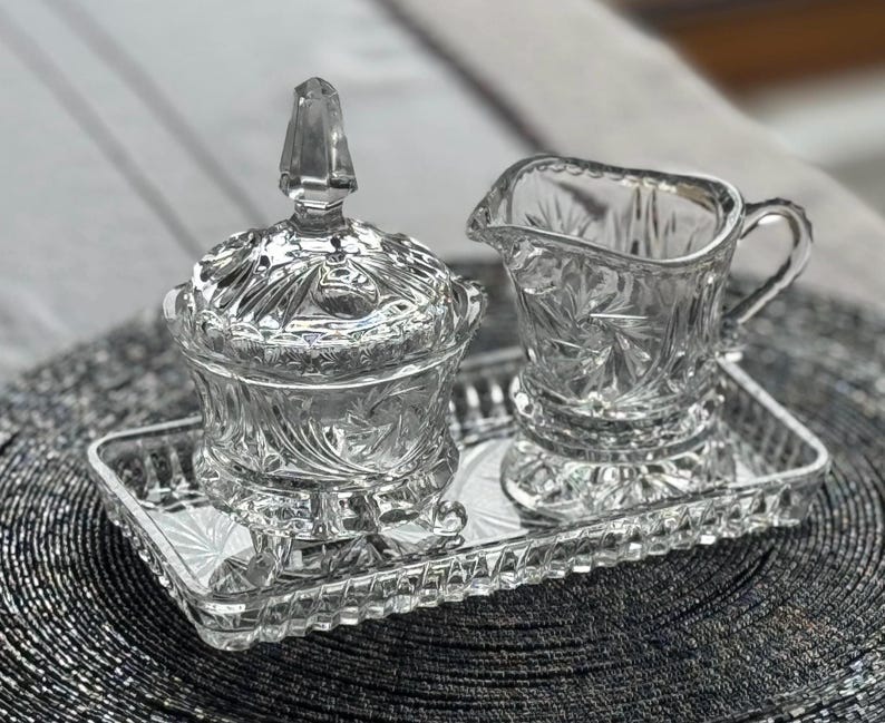 May include: A clear glass creamer and a lidded sugar bowl with a faceted finial, both featuring intricate starburst and geometric patterns, arranged together on a matching rectangular glass tray with a textured, scalloped edge design.