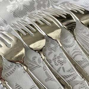 May include: A collection of silver forks arranged on a white patterned tablecloth. The forks have a classic design with tines and decorative handles. The silverware is likely for formal dining and adds elegance to any table setting.