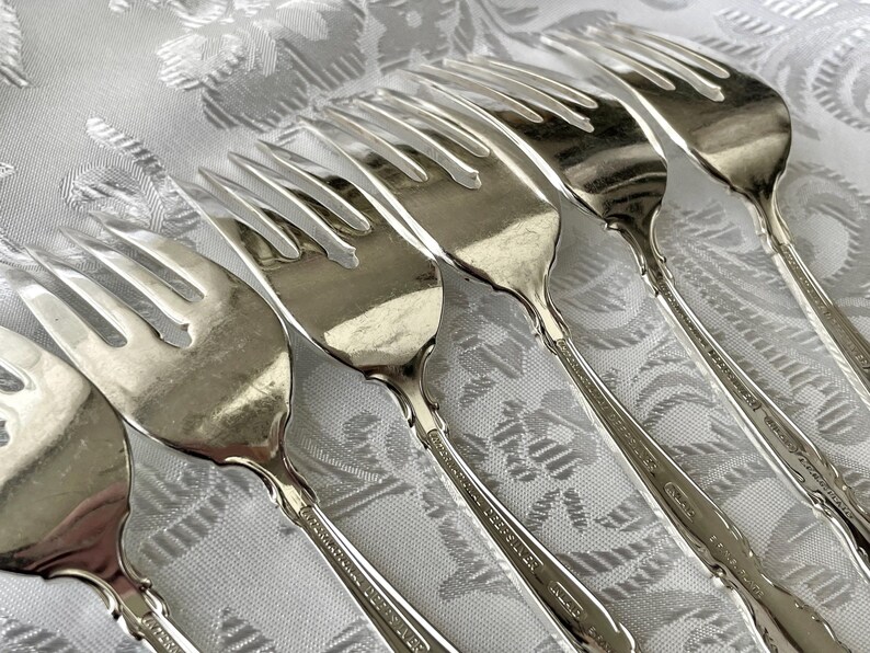 May include: A collection of silver forks arranged on a white patterned tablecloth. The forks have a classic design with tines and decorative handles. The silverware is likely for formal dining and adds elegance to any table setting.