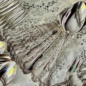 May include: A collection of ornate silver-colored flatware, including spoons and forks, arranged on a white lace surface. The silverware features detailed, decorative handles and reflective surfaces, suggesting a formal dining setting.