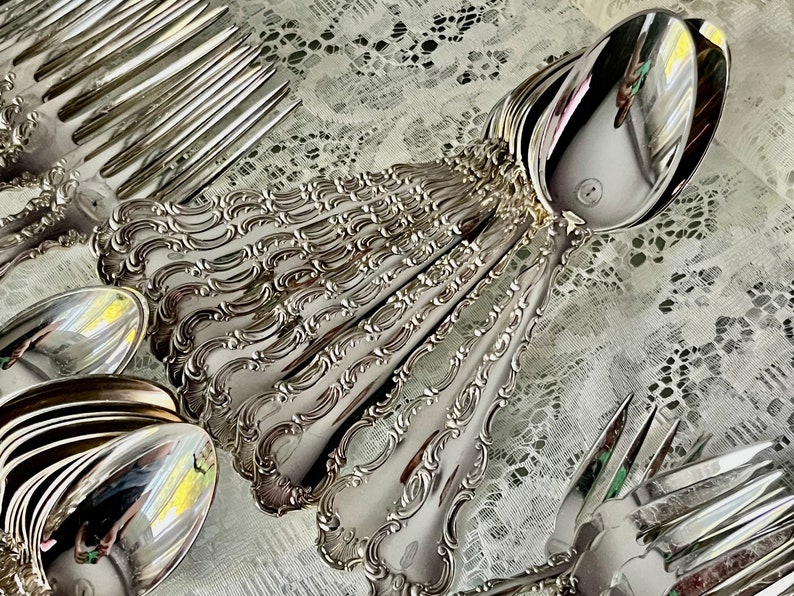 May include: A collection of ornate silver-colored flatware, including spoons and forks, arranged on a white lace surface. The silverware features detailed, decorative handles and reflective surfaces, suggesting a formal dining setting.