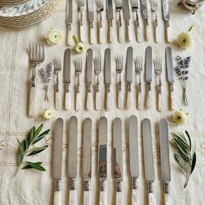 May include: An assortment of vintage silverware, including knives and forks, arranged on a cream-colored tablecloth. The silverware features mother-of-pearl handles and ornate silver detailing. Floral accents and decorative plates complete the elegant table setting.