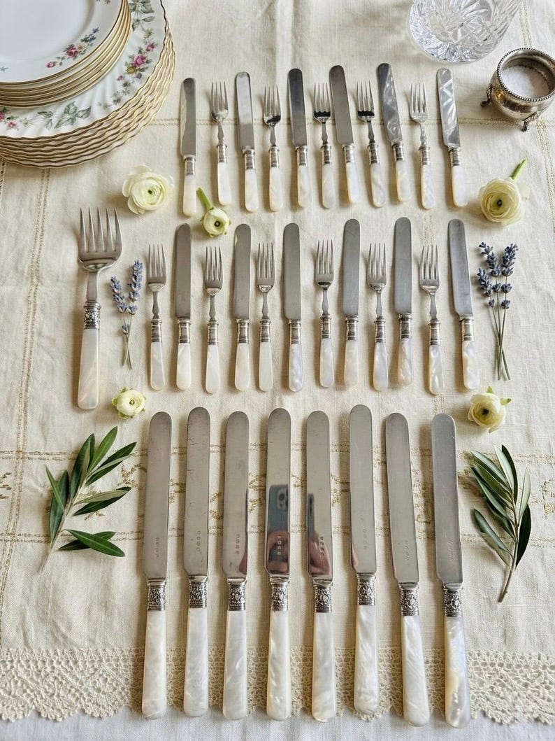 May include: An assortment of vintage silverware, including knives and forks, arranged on a cream-colored tablecloth. The silverware features mother-of-pearl handles and ornate silver detailing. Floral accents and decorative plates complete the elegant table setting.