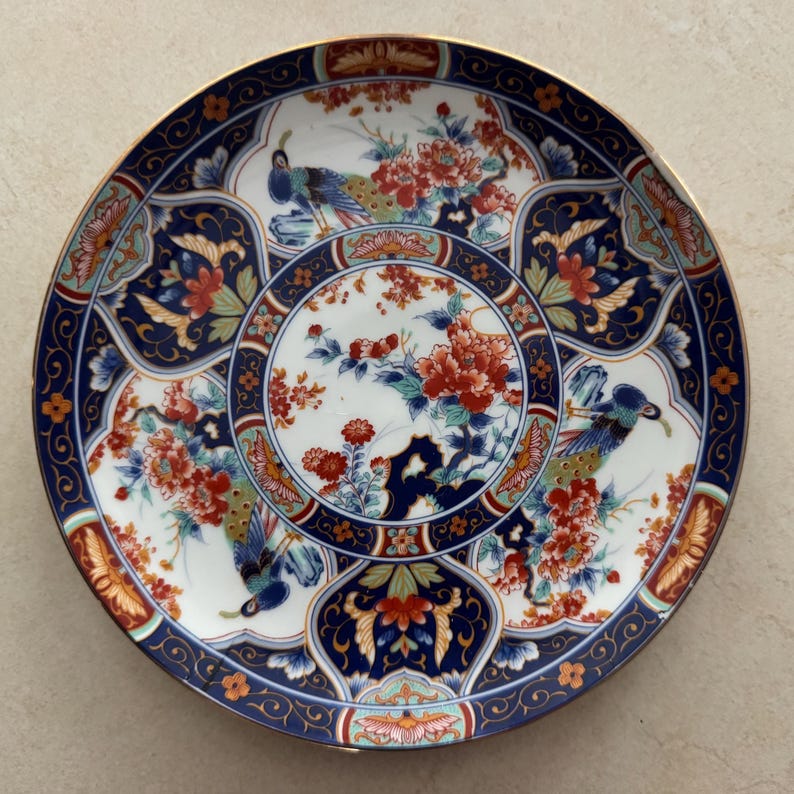 Pu&ograve; includere: Un piatto rotondo in porcellana con un medaglione circolare centrale con motivi floreali rossi e arancioni. Il bordo presenta uno sfondo blu intenso con intricati filigrana dorati, intervallati da illustrazioni di pavoni e motivi floreali.