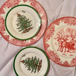 以下が含まれることがあります： 赤と緑の縁取りが施された白いセラミック製のクリスマス用プレート3枚。中央のプレートには、そりを引くトナカイの赤と白のイラストが描かれています。2枚の小さなプレートには、緑のクリスマスツリーが描かれています。