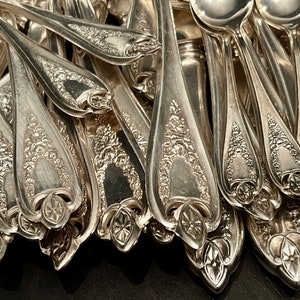 May include: A collection of silver-toned metal spoons featuring intricate embossed floral patterns on the handles. Each spoon handle displays a detailed decorative motif with a central geometric element at the base, showcasing a polished finish.