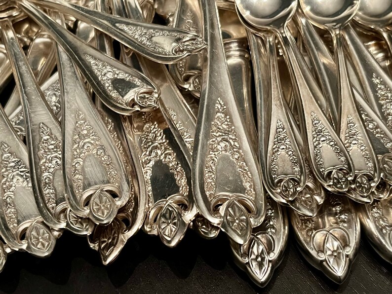 May include: A collection of silver-toned metal spoons featuring intricate embossed floral patterns on the handles. Each spoon handle displays a detailed decorative motif with a central geometric element at the base, showcasing a polished finish.