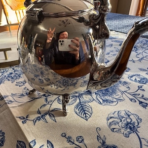 May include: A polished silver-plated teapot with a hinged lid, a curved spout, and three ornate feet. The reflective surface of the metal body shows a distorted reflection of a person holding a white smartphone while making a peace sign.