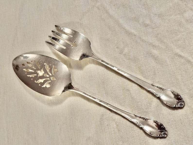 K&ouml;nnte beinhalten: Zwei silberne Servierbesteck, eines mit einem dekorativen Blumendesign und das andere mit einem gabelartigen Design.