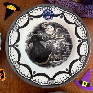 May include: A black and white Halloween themed dinner plate featuring a witch flying over a forest scene. The plate has a grey rim and a black and white border with a scalloped edge. The plate is made by Royal Stafford and says "Made in England".