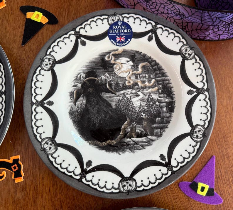 May include: A black and white Halloween themed dinner plate featuring a witch flying over a forest scene. The plate has a grey rim and a black and white border with a scalloped edge. The plate is made by Royal Stafford and says "Made in England".