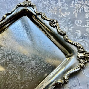 Puede incluir: Una bandeja rectangular de metal plateado con un intrincado patr&oacute;n floral grabado en su superficie. La bandeja tiene un borde elevado y ornamentado con bordes festoneados y motivos en forma de concha en las cuatro esquinas.