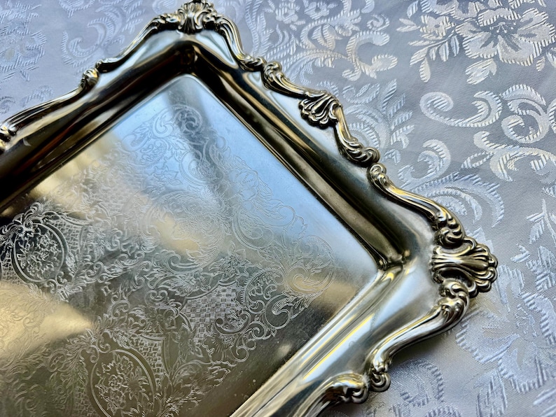 Puede incluir: Una bandeja rectangular de metal plateado con un intrincado patr&oacute;n floral grabado en su superficie. La bandeja tiene un borde elevado y ornamentado con bordes festoneados y motivos en forma de concha en las cuatro esquinas.