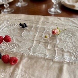 May include: A rectangular clear glass serving tray with a woven pattern and floral designs. The tray is decorated with raspberries, black currants, a small pink rose, and a daisy. The tray is on a cream-colored tablecloth.