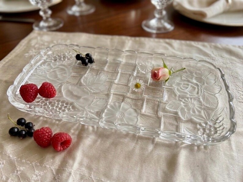 May include: A rectangular clear glass serving tray with a woven pattern and floral designs. The tray is decorated with raspberries, black currants, a small pink rose, and a daisy. The tray is on a cream-colored tablecloth.