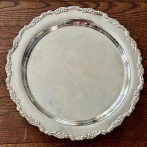 May include: A round silver serving tray featuring a polished center with an intricate, subtle floral engraving. The wide, scalloped rim is decorated with a detailed embossed pattern of small flowers and leaves along the entire edge.