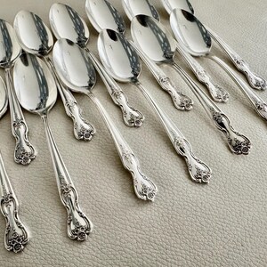 May include: A collection of silver spoons with ornate floral detailing on the handles. The spoons are arranged on a light-colored surface, reflecting the surrounding light. The spoons are a classic design, suitable for formal dining.