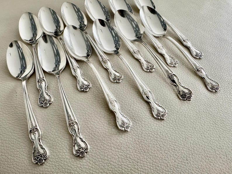 May include: A collection of silver spoons with ornate floral detailing on the handles. The spoons are arranged on a light-colored surface, reflecting the surrounding light. The spoons are a classic design, suitable for formal dining.