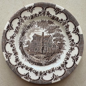 May include: A round ceramic plate featuring a dark brown transferware pattern on a cream background. The center depicts Little Morton Hall Cheshire with trees and a bridge. The rim is decorated with a floral rose vine and striped border.