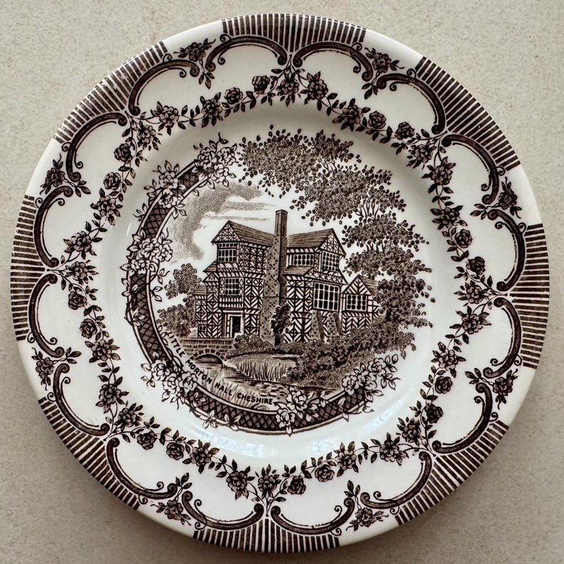 May include: A round ceramic plate featuring a dark brown transferware pattern on a cream background. The center depicts Little Morton Hall Cheshire with trees and a bridge. The rim is decorated with a floral rose vine and striped border.