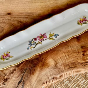 May include: A white ceramic dish with a wavy edge and gold trim. The dish is decorated with a floral pattern in shades of pink, yellow, blue, and green.
