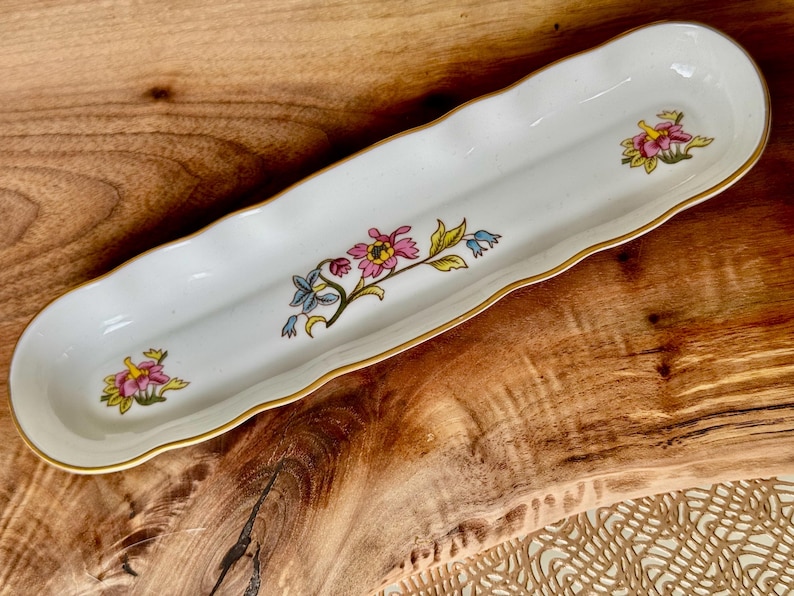 May include: A white ceramic dish with a wavy edge and gold trim. The dish is decorated with a floral pattern in shades of pink, yellow, blue, and green.