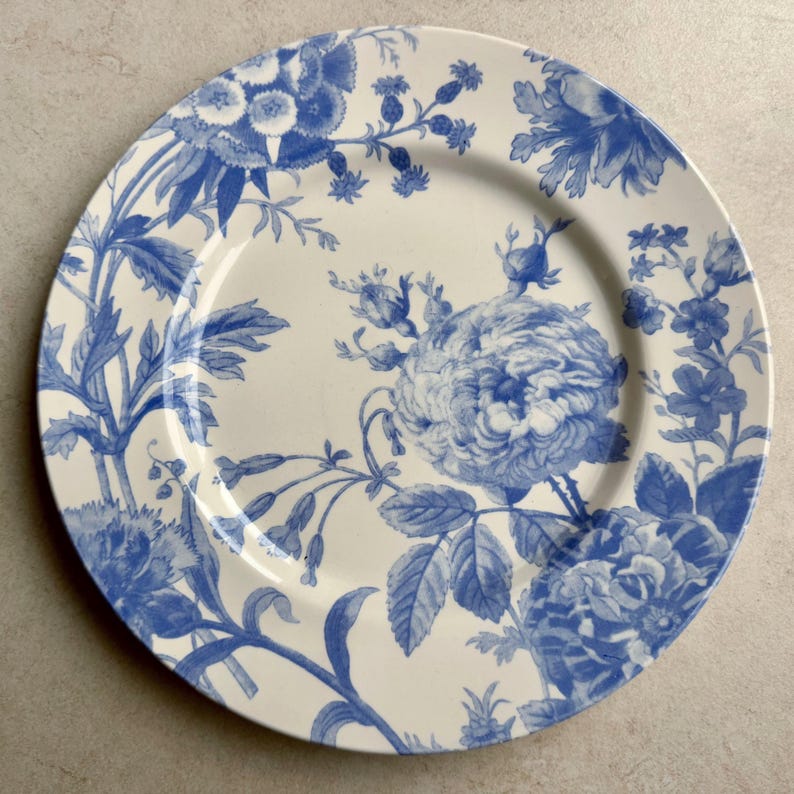 Puede incluir: Un plato redondo de cer&aacute;mica con base blanca y un patr&oacute;n floral azul. El dise&ntilde;o incluye ilustraciones detalladas de varias flores, capullos y hojas distribuidos por la superficie, creando un motivo bot&aacute;nico en tonos de color azul.