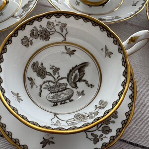 May include: A white teacup and saucer with a gold rim and black floral design. The design features a bird and a vase with a floral motif.