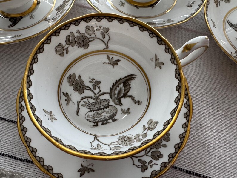 May include: A white teacup and saucer with a gold rim and black floral design. The design features a bird and a vase with a floral motif.