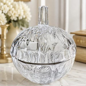 May include: A clear, round, cut-glass lidded bowl featuring a faceted, starburst-like pattern across its surface. The lid is topped with a vertical, ribbed handle, and the entire piece is crafted from transparent glass with reflective edges.