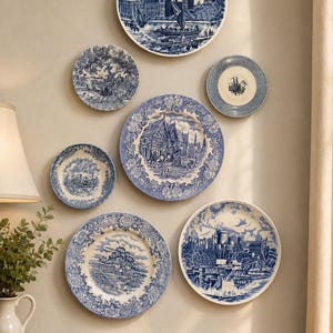 May include: A collection of six round ceramic plates displayed on a wall, each featuring blue and white transferware patterns. The designs include architectural landmarks, rural landscapes, and intricate floral borders on various plate sizes.