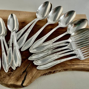 May include: A set of vintage silverware, including spoons and forks, arranged on a wooden cutting board. The silverware is made of polished silver and has a classic design.