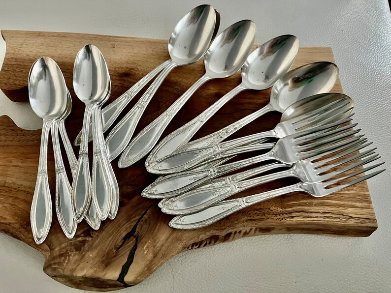May include: A set of vintage silverware, including spoons and forks, arranged on a wooden cutting board. The silverware is made of polished silver and has a classic design.