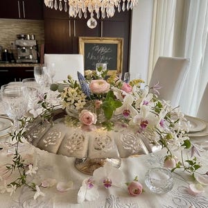 May include: A festive table setting with a silver cake stand centerpiece overflowing with fresh flowers, including orchids and roses. The table is set with crystal glasses and white plates, creating an elegant and celebratory atmosphere. A chalkboard with "Happy New Year" is in the background.