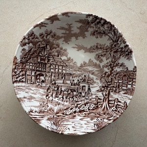 May include: A round ceramic bowl featuring a brown and white transferware pattern. The design depicts a horse-drawn carriage, timber-framed buildings, trees, and a stream. A sign on the building reads A WELCOME INN CO.