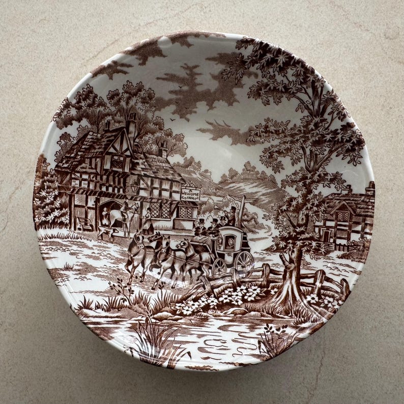 May include: A round ceramic bowl featuring a brown and white transferware pattern. The design depicts a horse-drawn carriage, timber-framed buildings, trees, and a stream. A sign on the building reads A WELCOME INN CO.