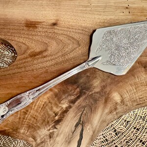 K&ouml;nnte beinhalten: Ein silberfarbener Tortenheber mit einem komplizierten, gepr&auml;gten Blumenmuster auf der dreieckigen Klinge und einem dekorativen, gewellten Design am Griff. Die Metalloberfl&auml;che ist poliert und reflektierend und zeigt detaillierte Schn&ouml;rkel.