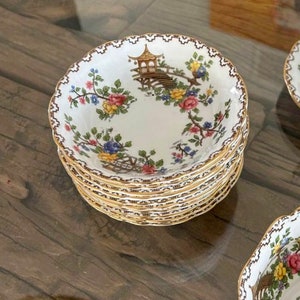K&ouml;nnte beinhalten: Ein Stapel wei&szlig;er Porzellanschalen mit goldfarbenem Rand und Blumenmuster. Die Schalen zeigen ein zartes Blumenmuster mit einem Pagoden- und Br&uuml;ckenmotiv.