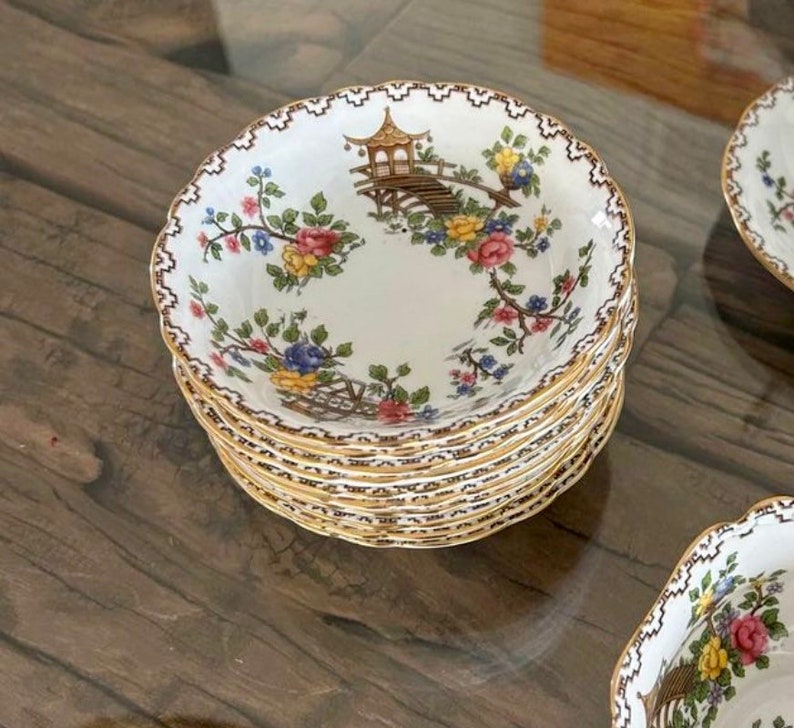 K&ouml;nnte beinhalten: Ein Stapel wei&szlig;er Porzellanschalen mit goldfarbenem Rand und Blumenmuster. Die Schalen zeigen ein zartes Blumenmuster mit einem Pagoden- und Br&uuml;ckenmotiv.