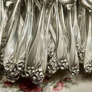 May include: A collection of ornate silver spoons and forks. The silverware features a floral design at the base of the handles. The spoons and forks are arranged closely together, reflecting light and creating a bright, shiny appearance. The overall theme is vintage tableware.
