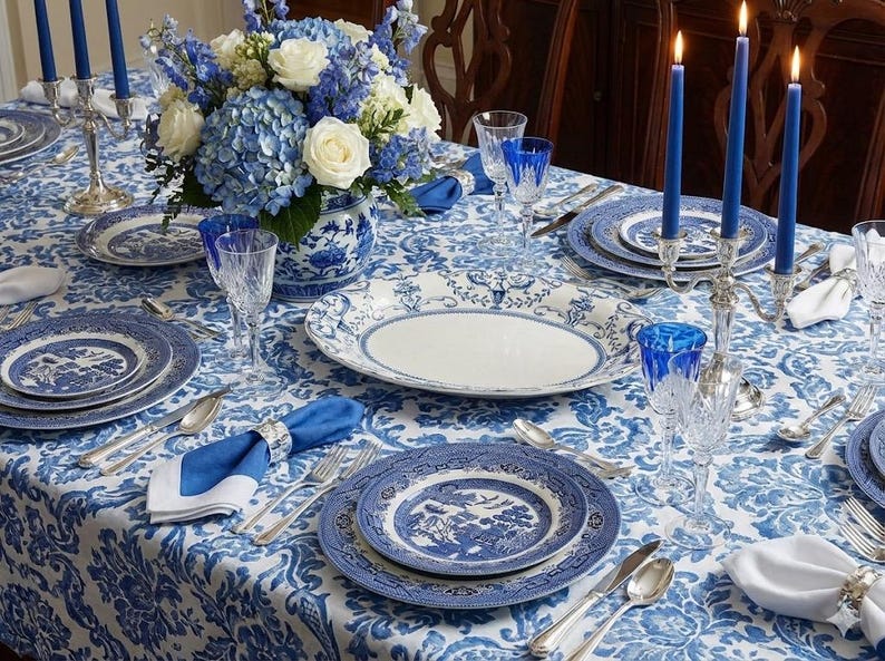 K&ouml;nnte beinhalten: Ein festlich gedeckter Esstisch mit blau-wei&szlig;em Porzellan, Kristallgl&auml;sern und Silberbesteck. Der Tisch ist mit einem Blumengesteck und brennenden Kerzen geschm&uuml;ckt, alles auf einer gemusterten Tischdecke. Eine klassische und elegante Einrichtung.