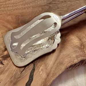 May include: Silver serving tongs with an ornate, engraved design. The tongs are open and resting on a wooden surface.