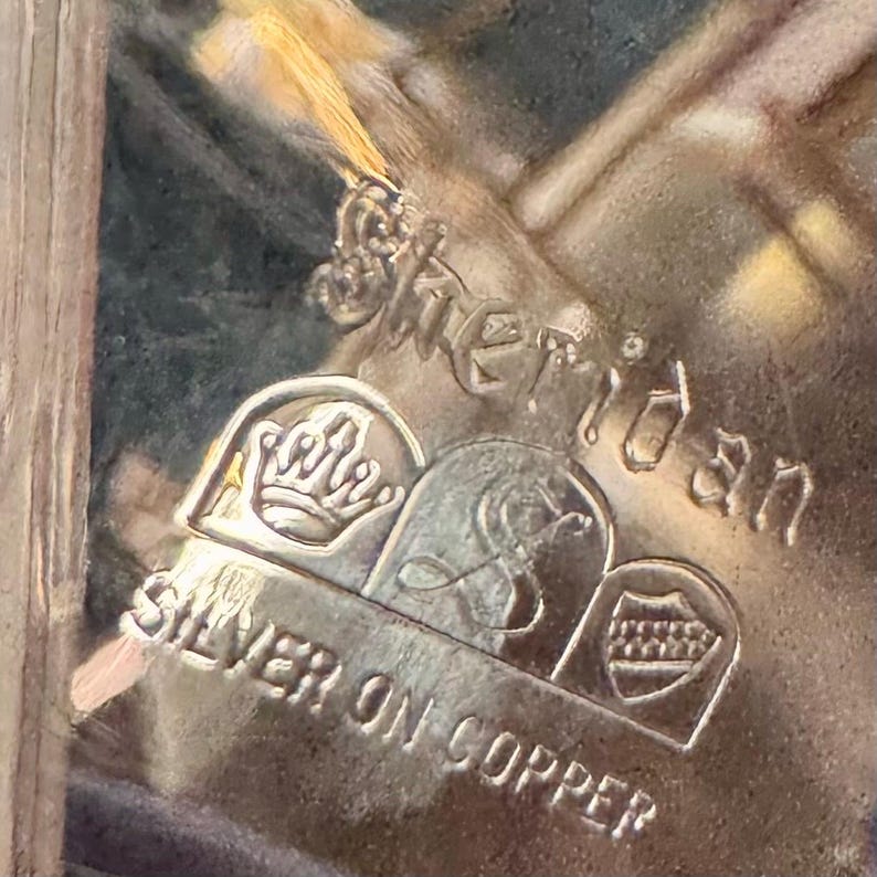 May include: A metallic maker's mark stamped on a surface, featuring the text Sheridan, Silver on Copper, and three distinct emblems: a crown, a stylized letter S, and a shield with horizontal lines, all embossed in a silver-toned metal.