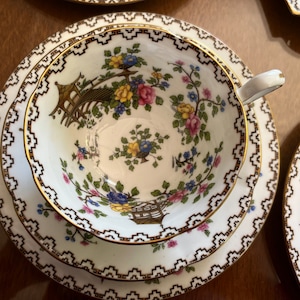 K&ouml;nnte beinhalten: Ein wei&szlig;es Tee-Set mit goldener Verzierung und einem floralen Muster. Das Muster zeigt eine Pagode, Blumen und V&ouml;gel. Die Untertasse hat einen doppelten Rand.