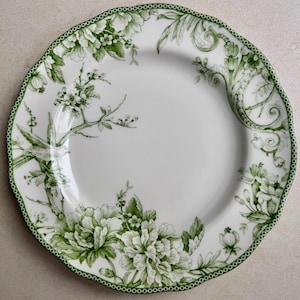 May include: A white ceramic plate with a scalloped edge and a green border featuring a repeating dot pattern. The plate is decorated with green botanical illustrations of blooming flowers, leafy branches, and a small bird perched on a twig.