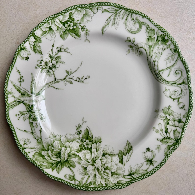 May include: A white ceramic plate with a scalloped edge and a green border featuring a repeating dot pattern. The plate is decorated with green botanical illustrations of blooming flowers, leafy branches, and a small bird perched on a twig.