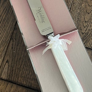 May include: A white cake knife with a silver blade and a white handle. The blade has a serrated edge and the handle has a floral design. The knife is in a pink box with the words "Wedding Memories" and "Stainless Steel Made in England" printed on it.