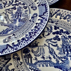 K&ouml;nnte beinhalten: Ein Stapel wei&szlig;er Keramikteller mit blau-wei&szlig;em Chinoiserie-Design. Die Teller zeigen eine detaillierte Landschaftsszene mit Pagoden, Br&uuml;cken und V&ouml;geln. Die R&auml;nder haben eine dekorative Bord&uuml;re. Die Teller sind auf einer Holzoberfl&auml;che gestapelt.