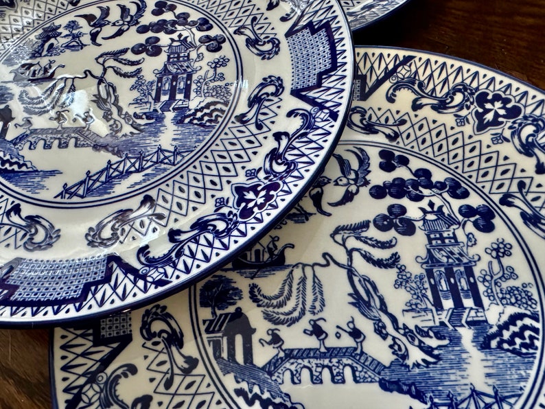 K&ouml;nnte beinhalten: Ein Stapel wei&szlig;er Keramikteller mit blau-wei&szlig;em Chinoiserie-Design. Die Teller zeigen eine detaillierte Landschaftsszene mit Pagoden, Br&uuml;cken und V&ouml;geln. Die R&auml;nder haben eine dekorative Bord&uuml;re. Die Teller sind auf einer Holzoberfl&auml;che gestapelt.
